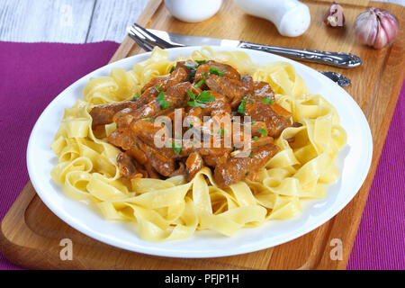 Pâtes fettuccine, garnie de délicieux bœuf stroganoff mijotée dans une sauce à la crème poêlée de champignons ceps sur plaque blanche sur une planche à découper avec cutle Banque D'Images