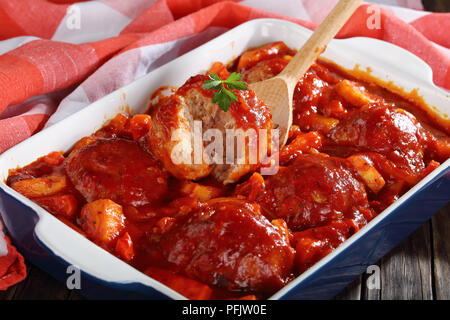 Close-up de cuillère en bois avec divisé en parties Meatball braisé à la sauce tomate et légumes au fond d'un plat allant au four avec des boulettes de viande sur sol en bois Banque D'Images