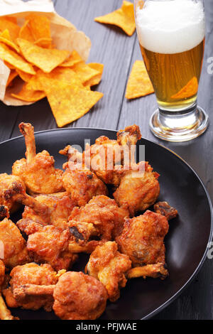 Frites délicieuses ailes de poulet croustillantes battues avec des os sur une plaque noire sur la table en bois sombre. verre de bière fraîche et des nachos en papier b Banque D'Images