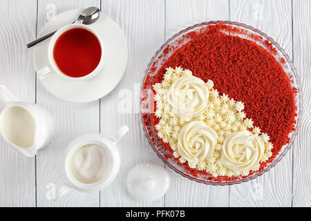 Délicieux gâteau de velours rouge surmonté de belles roses crème et parsemé de miettes de gâteau éponge rouge sur la plaque sur une table en bois blanc avec une tasse de thé, Banque D'Images