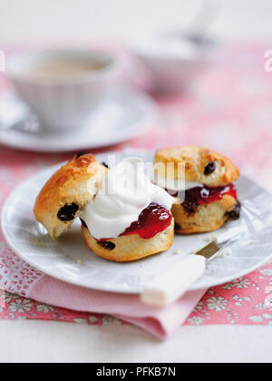 Scones aux raisins garni de confiture et crème fouettée, servi sur une assiette avec un couteau, et une tasse de thé en arrière-plan Banque D'Images