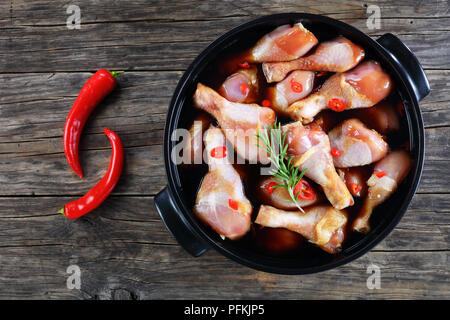 Pilons de poulet cru mariné à la sauce teriyaki épicé fait maison en noir pot sur table en bois foncé avec du chili à l'arrière-plan, Vue de dessus Banque D'Images