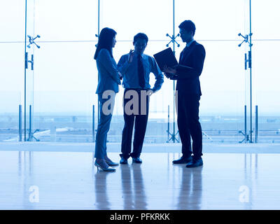 Trois personnes de l'entreprise asiatique debout devant la fenêtre de discuter affaires avec ordinateur portable. Banque D'Images