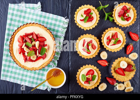 Tartelettes aux fraises et aux bananes, chargé avec crème pâtissière sur un plateau en ardoise noire, avec du sirop dans un bol sur la table en bois, voir f Banque D'Images