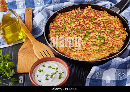 Une délicieuse cuisine suisse rosti ou galette de pommes de terre saupoudrées de persil finement haché dans une poêle sur une table en bois noir avec de la crème dans un bol, close-up Banque D'Images