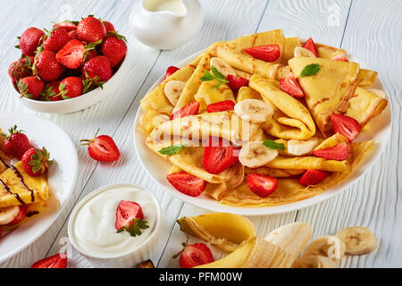 Crêpes minces ou des crêpes sur une plaque avec des fraises, des tranches de banane et de feuilles de menthe, la crème et le bol avec des fraises fraîches à l'arrière-plan, ho Banque D'Images