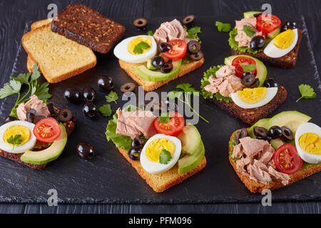 Sandwiches au thon avec des tranches d'avocat, salade, tomates, olives noires et oeuf dur sur une tranche de pain de seigle et de maïs les tranches de bac en ardoise noire Banque D'Images