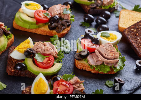 Close-up de sandwiches au thon avec des tranches d'avocat, salade, tomates, olives noires et oeuf dur sur le seigle et le maïs des tranches de pain grillé sur une black s Banque D'Images