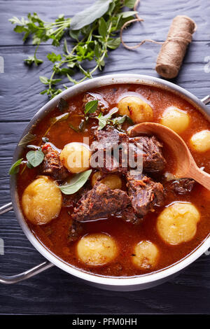 Ragoût de boeuf avec des cubes de viande tendre, ensemble de nouvelles pommes de terre, carottes et herbes dans un chaudron sur une table en bois noir avec bouquet d'herbes aromatiques, vertic Banque D'Images