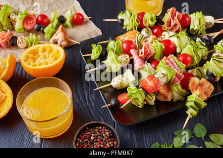 L'antipasto brochettes avec la viande de poulet, courgettes, tomates, mozzarella tranches de salami, boules, des olives sur une plaque noire sur une table en bois avec du jus d'orange Banque D'Images