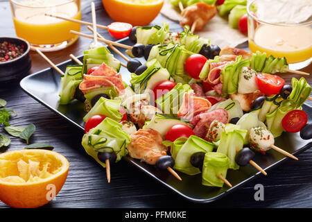 Kebab en brochettes avec la viande de poulet, courgettes, tomates, mozzarella tranches de salami, boules, des olives sur une plaque noire sur une table en bois avec fresh orange j Banque D'Images