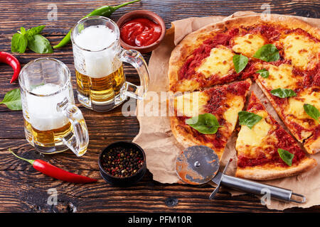 Close-up of classic Italian Pizza Margherita coupé en tranches sur un papier parchemin sur une vieille table en bois rustique avec de la bière dans des tasses en verre, vue de abov Banque D'Images