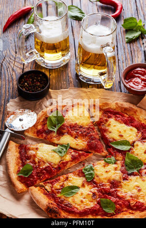 Classic Italian Pizza Margherita coupé en tranches sur un papier parchemin sur une vieille table en bois rustique avec de la bière dans des tasses en verre, recette authentique, vertical Banque D'Images