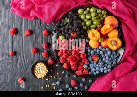 Salade de fruits et baies avec des produits bio frais de groseille, framboise rouge et noire, de bleuet, de tranches d'abricots sur une plaque noire sur une table en bois avec des pins Banque D'Images