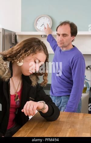 Fille assise à une table de cuisine à sa montre, l'homme en montrant une horloge Banque D'Images