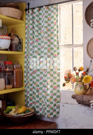 Fenêtre chalet vu de l'intérieur ; rideau oeillet avec crochet vert et des ustensiles de cuisine sur la pendaison de fer sur fenêtre, vase de fleurs sur table en bois rugueux, jaune à côté des étagères. Banque D'Images