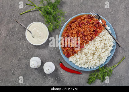 Close-up of sex délicieux chili con carne avec des haricots rouges servi sur une assiette avec du riz bouilli sur table béton avec de la crème sure, vue d'en haut Banque D'Images