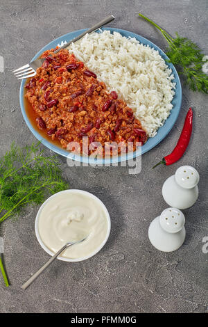 Sex délicieux chili con carne avec des haricots rouges servi sur une assiette avec du riz bouilli sur table béton avec de la crème sure, vue verticale d'en haut Banque D'Images