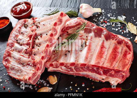 Carré de porc frais crus de côtes assaisonnées d'épices sur le plateau en ardoise noire avec le paprika, l'ail, les feuilles de laurier à l'arrière-plan, vue verticale d'un Banque D'Images