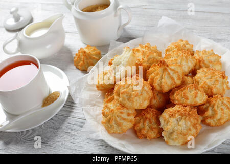 Des délicieux cookies à la noix de coco sur une plaque blanche sur table en bois avec tasse de thé, la cuvette et la crème fraîche dans un pot à lait à l'arrière-plan, vue depuis Banque D'Images