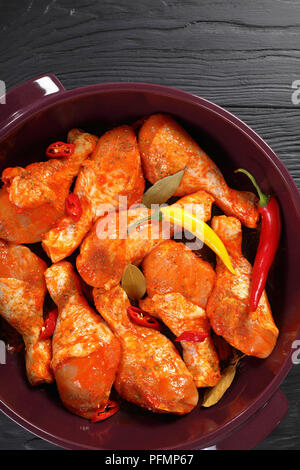 Pilons de poulet cru mariné frotté avec la spice, morceaux de piment, les feuilles de laurier prêt à cuire dans un plat, vue verticale de abov Banque D'Images
