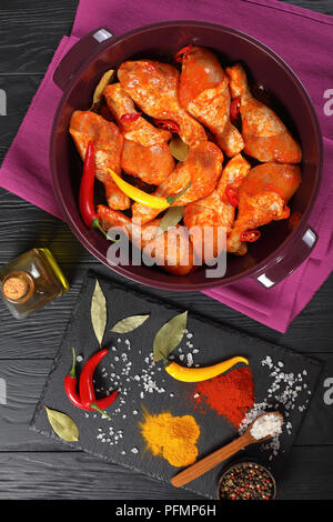 Pilons de poulet cru mariné frotté avec la spice, morceaux de piment, les feuilles de laurier prêt à cuire dans un plat, les ingrédients sur une pierre Banque D'Images