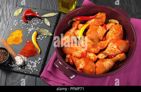 Pilons de poulet cru mariné saupoudré d'épices, morceaux de piment, les feuilles de laurier prêt à cuire dans un plat, les ingrédients sur une carte sur une ardoise Banque D'Images