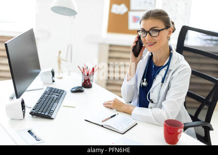 Une jeune fille dans une robe blanche est assis à une table dans le bureau et parle au téléphone. Un stéthoscope pend autour de son cou. Banque D'Images