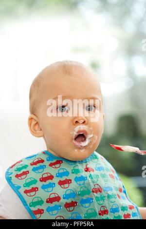 Baby Boy avec enduit alimentaire autour de sa bouche, le port d'un dossard, cuillère à proximité, looking at camera Banque D'Images