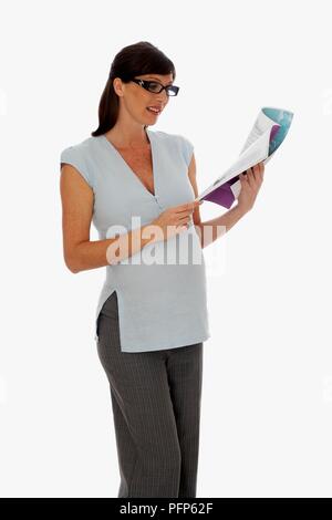 Femme enceinte en haut à manches courtes et un pantalon, le port de lunettes, à la recherche aux pages d'un magazine Banque D'Images