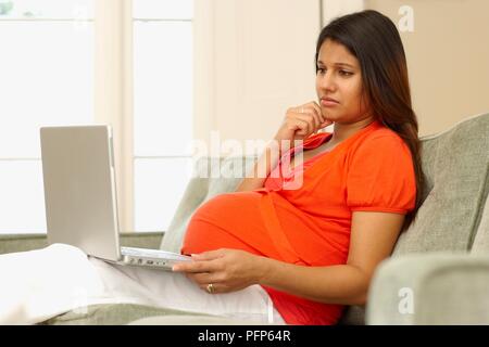 Pregnant woman sitting on sofa looking at laptop écran, side view Banque D'Images