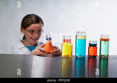 Pré-adolescente in lab coat, souriant, l'examen de l'un des cinq petites bouteilles contenant des liquides colorés sur la table Banque D'Images