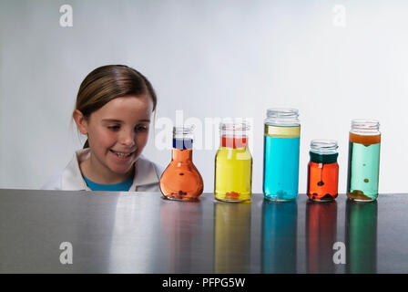 Pré-adolescente in lab coat, souriant, d'un examen de cinq petites bouteilles contenant des liquides colorés sur la table Banque D'Images