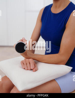 Femme portant gilet bleu top et short bleu clair assis sur une chaise, un coussin sur ses genoux, les bras croisés, poids levé dans une main, paume vers le haut, le poignet tordu vers le haut, close-up Banque D'Images