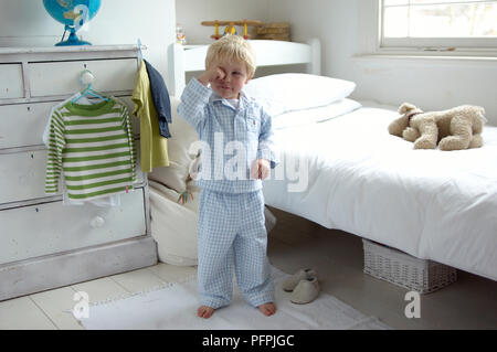 Boy wearing pyjamas, debout à côté de lit, se frottant les yeux Banque D'Images