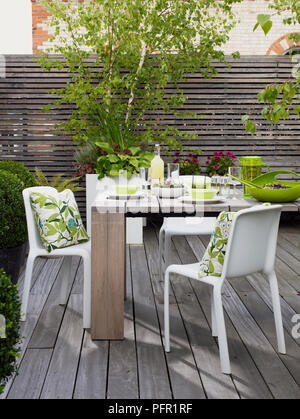 Set de table en bois pour manger vu de l'avant et coussins motif floral sur des chaises en plastique blanc sur le patio en bois Banque D'Images