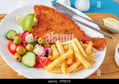 Le poisson frit à la dans la chapelure, servi avec des légumes frais, feta, olives salade grecque et les frites sur une plaque blanche avec fourchette et couteau d'argent sur Banque D'Images