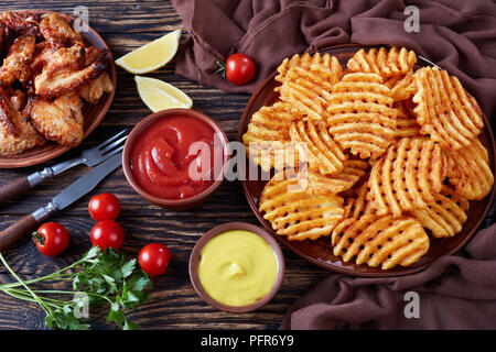 De pommes de terre frites croustillantes criss cross sur une plaque d'argile sur une table en bois avec de la moutarde et de la sauce tomate et tremper les ailes de poulet collant à l'arrière-plan, v Banque D'Images