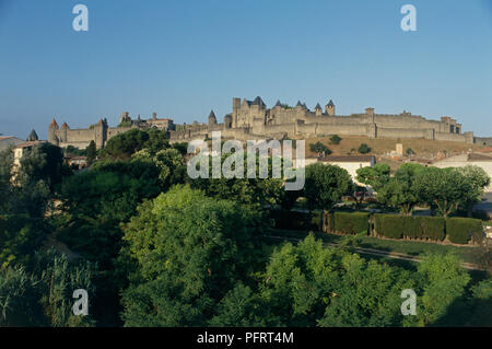 France, Aude, Cité de Carcassonne, ville médiévale fortifiée de murs à distance définie contre ciel bleu clair avec des arbres en premier plan Banque D'Images