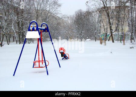 Aire enfantin recouvert de neige en hiver. Seul swing sur aire de jeux pour enfants au cours de la neige cyclone. Cour vide dans snowfal ville européenne après Banque D'Images