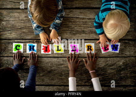 Vue de dessus de quatre enfants de races mixtes de toucher un mot écrit avec enfants lettres colorées sur cartes blanches plus de texture rustique les planches de bois. Banque D'Images