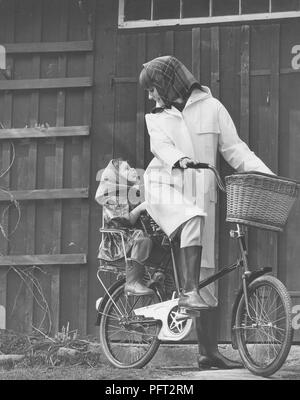 La pluie dans les années 1960. Une jeune femme souriante est habillé en vêtements de pluie et des manèges un vélo avec sa fille. Suède 1967 Banque D'Images