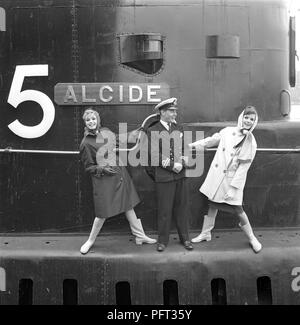 La mode des années 1960. Deux modèles de mode et le capitaine du sous-marin britannique Alcide. Le sous-marin est ancrée dans le port de Stockholm. Les modèles portent les automnes de mode imperméables et les foulards de tête. La Suède Mai 1962 Banque D'Images