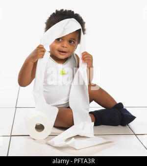 Boy wearing white vest et la marine chaussons assis sur marbre avec du papier toilette sur la tête, 17 mois Banque D'Images