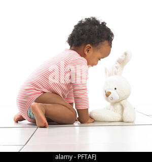 Fille à genoux vers un jouet lapin, 18 mois Banque D'Images