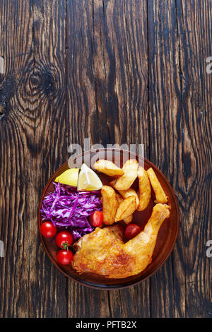 Quartiers de pommes de terre frites, cuisse de poulet et salade de chou frais servis sur une plaque d'argile sur une table en bois foncé Banque D'Images