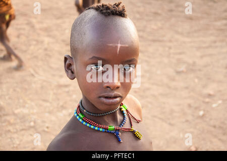Beau jeune garçon africain avec colliers traditionnels de mystérieux signe en forme de croix sur le front de la tribu Hamar à curieusement avec de magnifiques yeux brillants Banque D'Images