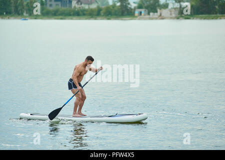 Weda de sportsman la natation dans le lac ville debout sur le conseil sup , laissant longue rame. Modèle masculin ayant fit figure, corps sportif, portant des shorts noir. Le maintien d'un mode de vie sain. Banque D'Images