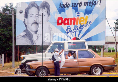 Managua, Nicaragua, élections, Feb 1990;adolescents vente de fruits, de tissus, de chemises et d'autres biens aux conducteurs à un carrefour avec Ortega affiche électorale derrière. Banque D'Images