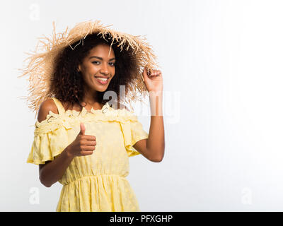 Jeune femme noire africaine giving thumb up sur fond blanc. Fille en chapeau de paille et robe jaune. Copier l'espace. Banque D'Images